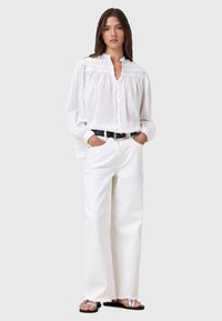 Blusa bianca in lino, morbida e con dettagli plissettati, chiusura con bottoni e maniche lunghe. Abbinata a jeans bianchi a gamba larga e sandali neri.