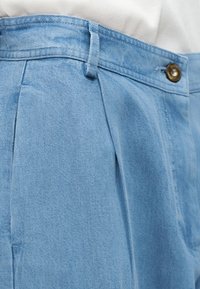Pantalon en denim bleu clair avec plis, passants pour ceinture et fermeture à bouton en laiton, porté avec un haut blanc rentré.