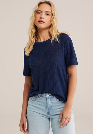 Femeie cu păr lung blond, purtând un tricou texturat cu mâneci scurte, bleumarin, și blugi albastru deschis, pe un fundal bej simplu.