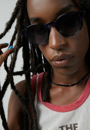 Lunettes de soleil rondes et sombres en matériau translucide bleu marine. Un collier ras du cou en perles noires est visible. Cheveux en longues tresses, teint clair.