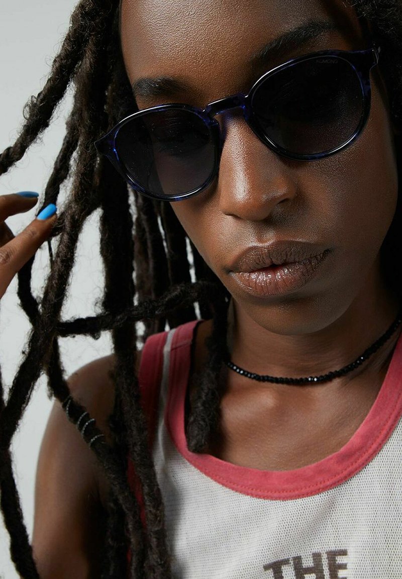 Ronde, donkere zonnebril gemaakt van doorschijnend marineblauw materiaal. Zichtbare zwarte kralen chokersieraden. Haar in lange, gevlochten lokken, lichte huidskleur.