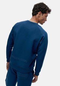 Temno modra majica s kapuco iz mehkega materiala z rebrastimi manšetami in robom, ki ima tekstovni detajl na spodnjem delu hrbta. Gladka tekstura, sproščen kroj.