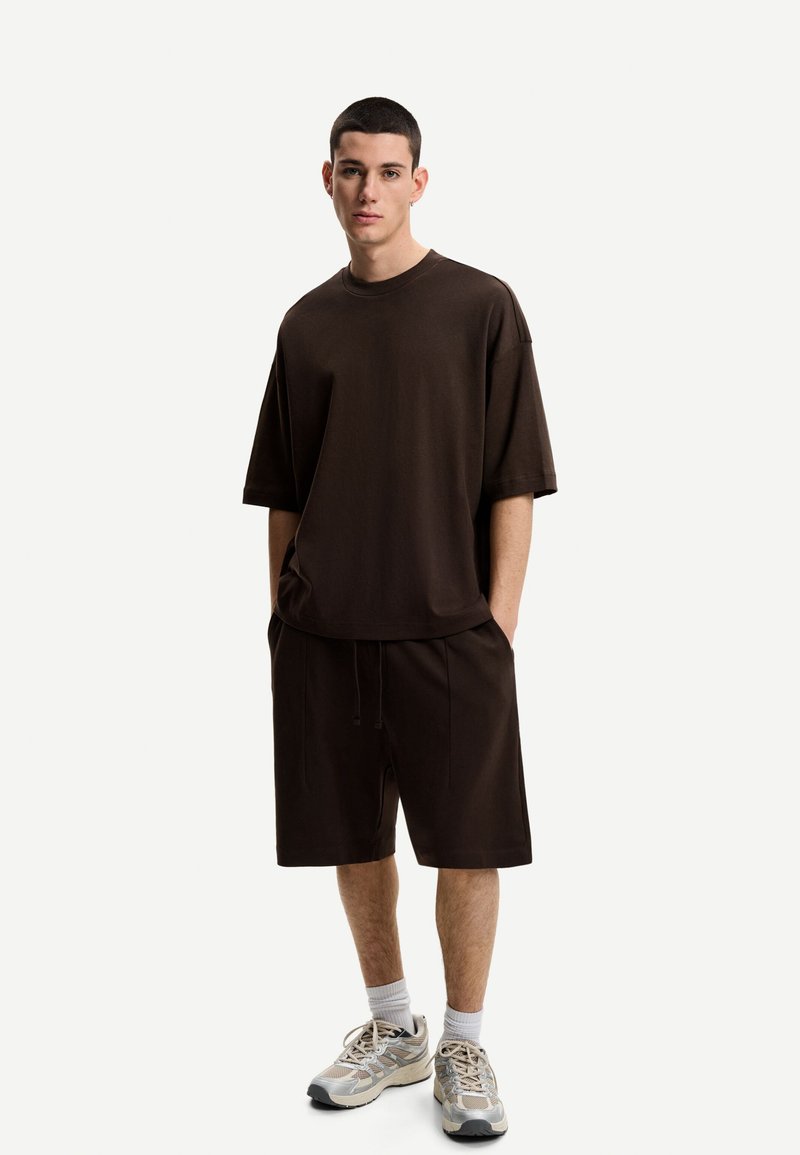 Hnedý bavlnený oversized tričko s krátkymi rukávmi, spárované s vyťahovacími šortkami v rovnakých farbách, doplnené viacfarebnými športovými teniskami.
