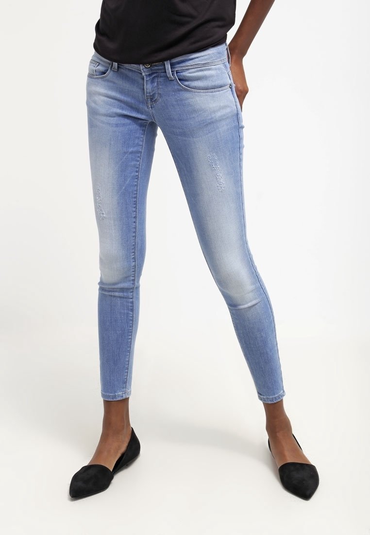 Persona vestida con pantalones ajustados de mezclilla azul claro desgastados, flats negros de punta y una blusa negra, de pie contra un fondo blanco liso.
