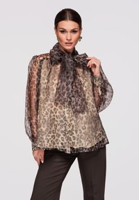 Femme portant un chemisier transparent à imprimé léopard avec un grand nœud au col et un pantalon foncé, posant devant un fond uni clair.