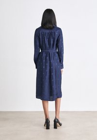 Vestito blu navy a maniche lunghe con una trama decorativa, cintura in vita e taglio a metà gamba; abbinato a scarpe con tacco alto nere.