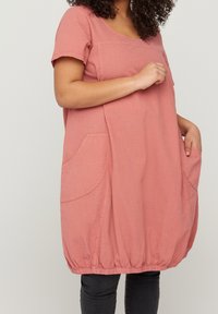 Kurvig kvinna bär en löst sittande rosa tunikaklänning med stora fickor, står mot en neutral bakgrund, händerna vilande på höfterna.