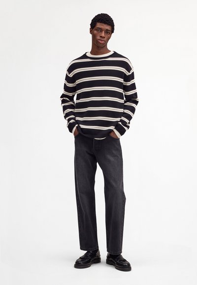 Nuori mies kädet taskuissa, yllään mustavalkoraidallinen neulepaita, mustat farkut ja mustat nahkakengät valkoista taustaa vasten.