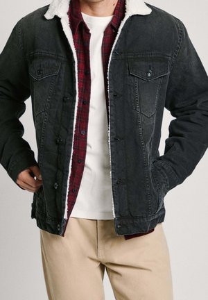 Mand iført beige bukser, hvid T-shirt, rød flannel-skjorte og sort denimjakke med hvid fleece-krave, hænderne i lommerne.