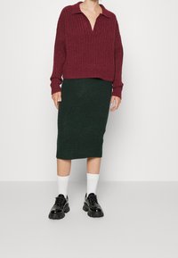 Burgundy stickad oversized tröja med krage, kombinerad med en mörkgrön ribbad midilång kjol och klumpiga svarta skor med vita strumpor.