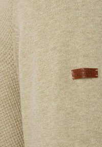 Beżowy sweter z dzianiny o wzorze teksturowym, z brązową skórzaną metką z napisem "Authentic Clothing REDMOND" naszytą na nim.