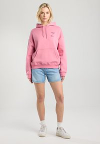 Pink hoodie made of cotton blend with a kangaroo pocket, ribbed cuffs, and hem; worn with light blue denim shorts and white sneakers.