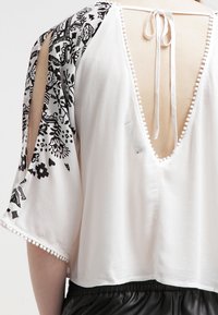 Blouse blanche avec manches noires à motifs, dos ouvert et fermeture à nouer ; ornée de petits pompons blancs le long de l'ourlet.
