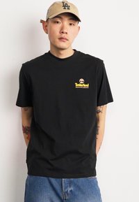 Camiseta de algodón negra con cuello redondo y logo de Timberland en amarillo en el lado izquierdo del pecho. Llevada con jeans de mezclilla azul claro.