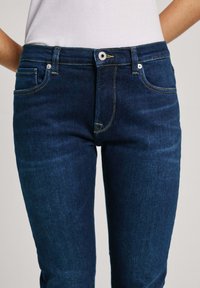 Jeans i denim med en mörkblå färg, med slim fit, femfickorsdesign, gula sömmar, knappknäppning framtill och metallbeslag.