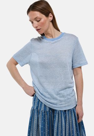 Femme regardant vers le bas portant une chemise à manches courtes rayée bleu clair et blanc et une jupe bleue à motifs, main sur la hanche.