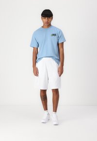 Light blue cotton t-shirt with small graphic on the chest, paired with white mesh athletic shorts, white sneakers, and visible leg tattoo.