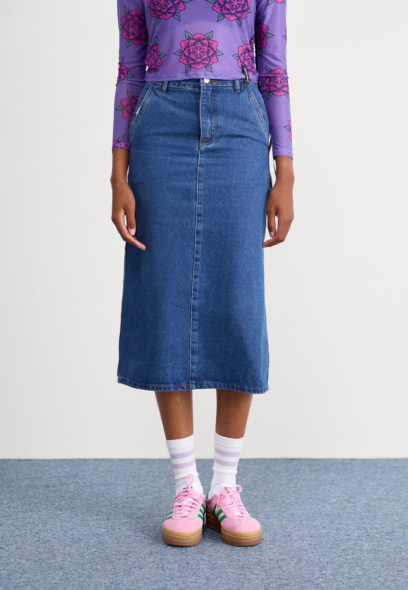 Jupe en denim de coupe droite, longueur mi-mollet, avec une couture frontale. Associée à des baskets roses et des chaussettes rayées blanches.