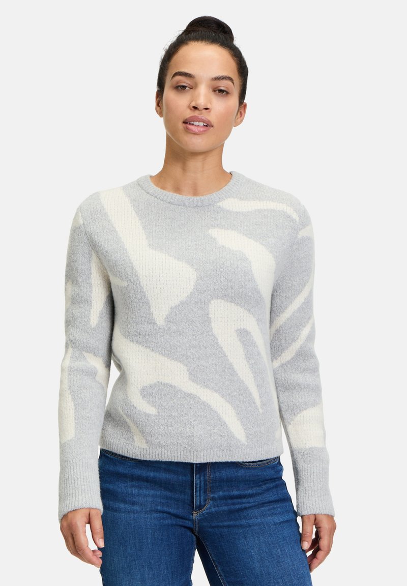 Hellgrauer Pullover mit einer weichen, strukturierten Oberfläche. Verfügt über weiße abstrakte Muster, einen gerippten Rundhalsausschnitt und lange Ärmel. Kombiniert mit blauen Jeans.