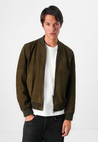 Καφέ σουέντ μπόμπερ τζάκετ με ριγωτό γιακά και μανίκια, κλείσιμο με φερμουάρ και δύο πλαϊνές τσέπες. Φορεμένο πάνω από λευκό t-shirt και μαύρα τζιν.