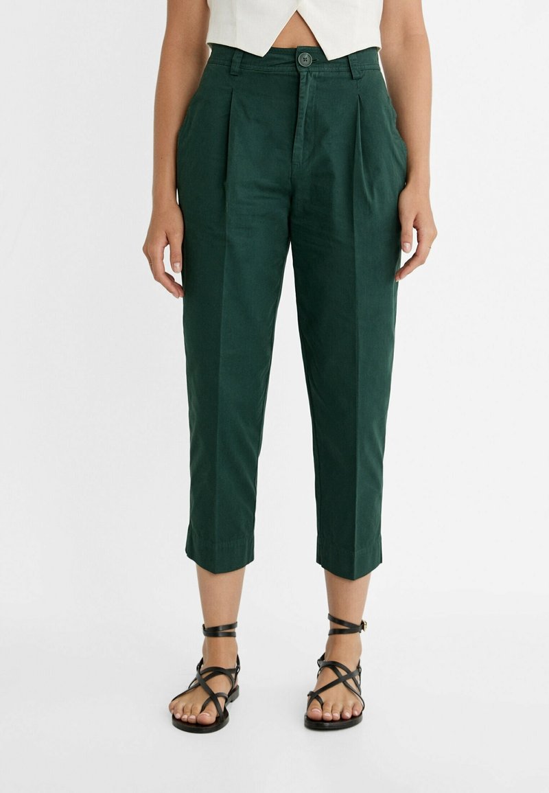 Femme portant un pantalon court taille haute vert foncé avec plis devant et des sandales noires à brides, debout devant un fond blanc.