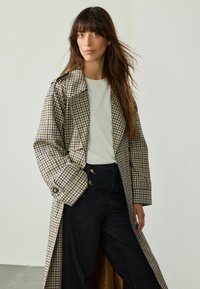 Beżowy i granatowy trench coat w kratę, z szerokim kołnierzem, dużymi guzikami oraz luźnym krojem, w zestawieniu z ciemnymi dżinsami.