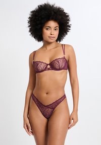 Ensemble lingerie en dentelle violet profond comprenant un soutien-gorge armatures orné de motifs floraux et un tanga assorti avec des détails similaires. Bretelles réglables.
