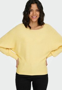 Żółty sweter w paski z szerokim dekoltem, o luźnym kroju i szerokich rękawach. Struktura tkaniny wydaje się miękka i prążkowana. Połączony z czarnymi dżinsami.