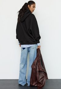 Černá oversized mikina s texturovaným povrchem, doplněná světlou modrou pruhovanou košilí a vybledlými širokými džínami, držíc hnědou koženou bundu.