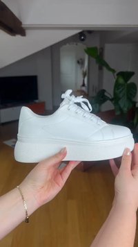 Handen houden een witte platform sneaker met een dikke, gestructureerde zool vast in een lichte binnenruimte met planten en meubels op de achtergrond.