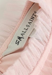 Notranjost svetloroza obleke s puhastimi volančki iz mehke tule. Oznaka z napisom "ALLSAINTS" je natisnjena črno in vidna na teksturirani tkanini.