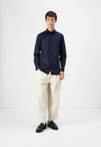 Camisa de botones azul marino con cuello, ajuste estilizado y mangas largas, combinada con pantalones anchos color crema. Mocasines negros completan el look.