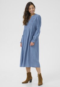 Denim mėlynos spalvos ilgos rankovės suknelė su tekstūruotu smukimo dekoru per krūtinę ir laisvu sijonu. Sunešiota su rudais zomšiniais auliniais batais iki kulkšnies.
