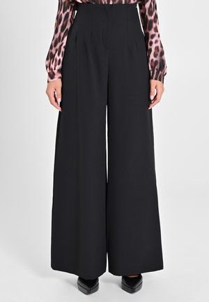 Pantalon noir à jambes larges en tissu lisse. Doté d'une taille haute avec des plis et d'une silhouette fluide. Porté avec des chaussures noires.