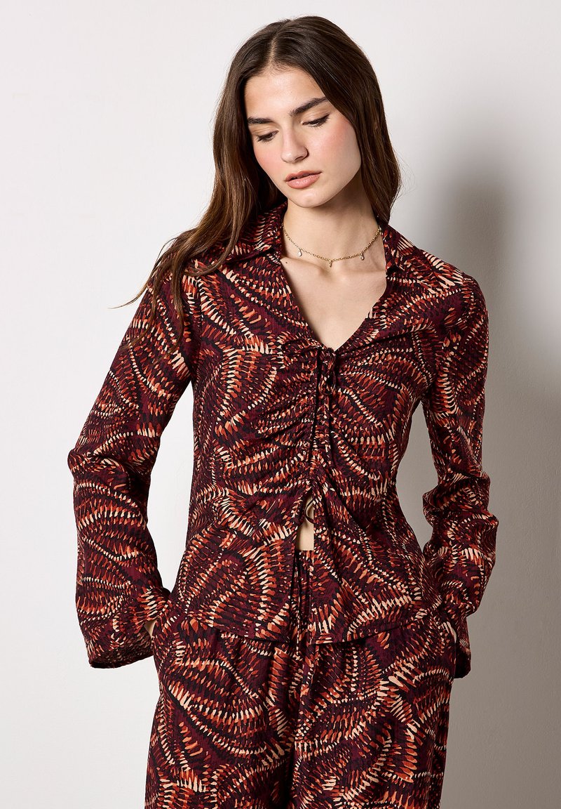 Mujer con cabello largo y castaño, vestida con un conjunto a juego de top y pantalones de manga larga con estampado en rojo, naranja y negro, de pie con las manos en los bolsillos.