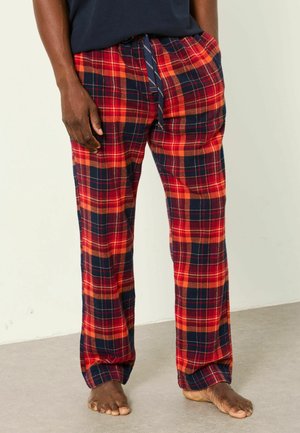 Man som bär röda, orange och marinblå rutiga pyjamasbyxor med marinblå dragsko och barfota står på ett ljust golv mot en vit vägg.