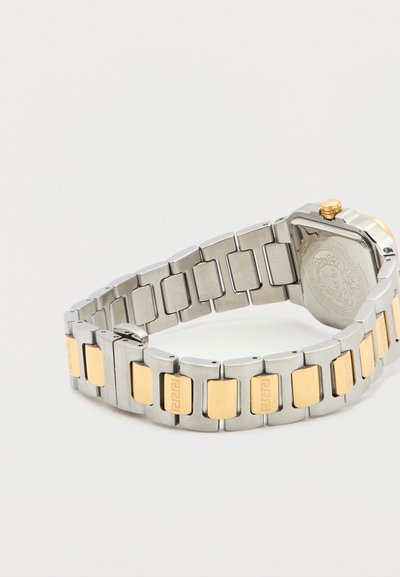 Μεταλλικό λουράκι ρολογιού με ασημένιους κρίκους και ορθογώνιες χρυσές πινελιές, που δείχνει την πίσω όψη της θήκης του ρολογιού και το χρυσό κουμπί σε γκρι φόντο.