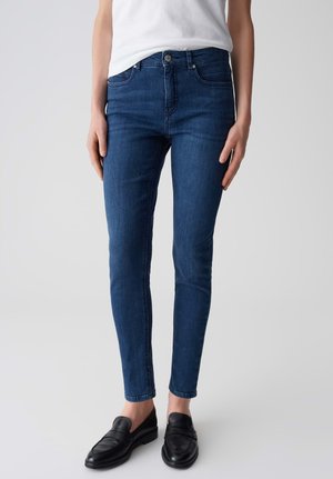 Jeans attillati di colore blu scuro con una consistenza liscia, dotati di chiusura con zip, design a cinque tasche e dettagli di cucitura sottili.