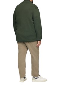 Grüner gerippter Strickpullover mit kragenloser Halslinie, kombiniert mit beigen Hosen und weißen Sneakern. Sichtbare Nähte und lockere Passform.