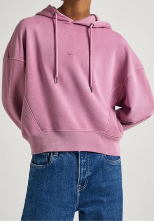 Personne portant un sweat à capuche rose ample avec cordons, associé à un jean en denim bleu taille haute sur un fond uni.