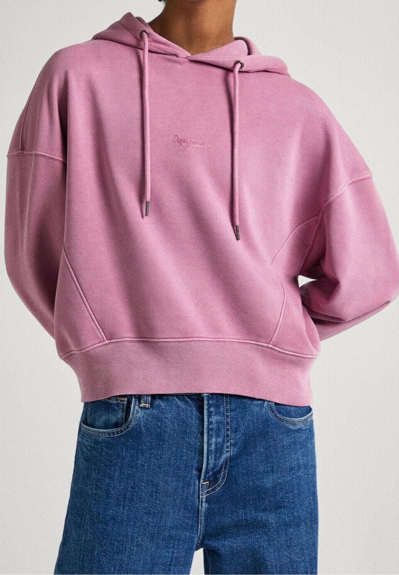 Personne portant un sweat à capuche rose ample avec cordons, associé à un jean en denim bleu taille haute sur un fond uni.