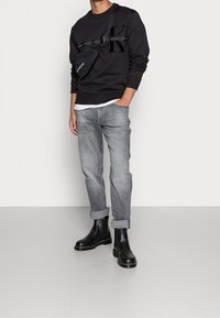Felpa nera con grande logo, jeans grigi e stivaletti neri alla caviglia. L'outfit include una borsa a tracolla scura. Design semplice e casual.