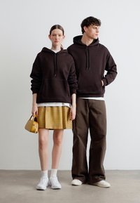 Μια γυναίκα με καφέ φούτερ με κουκούλα, κίτρινη καρό φούστα και λευκά αθλητικά παπούτσια κρατά μια κίτρινη τσάντα, δίπλα σε έναν άνδρα με καφέ φούτερ με κουκούλα και χαλαρό καφέ παντελόνι.