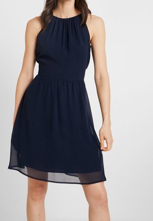 Mujer con un vestido sin mangas azul marino hasta la rodilla, con escote fruncido y falda de capas ligeramente transparente, posando frente a un fondo liso.