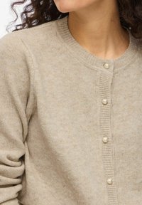 Smėlio spalvos megztinis kardiganas su apvaliu kaklu, ribotais kraštais ir perlamutriniais mygtukais priekyje. Minkšta tekstūra visame gaminyje.