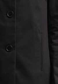 Lähikuva mustast kangast kostüümist, kus on kaks musta nööpi ja vertikaalne küljetasku, kus on nähtavad õmblused.