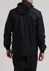 Mies, joka käyttää mustaa hupparia ja mustia housuja, seisoo selin kameraan neutraalilla taustalla.