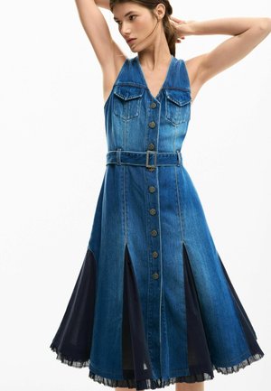 Naine kannab varrukateta sinist teksakleiti, millel on ees nupud, vööga talje ja mustad kangasissekanded lehvikulises seelikus, poseerides käed tõstetud.