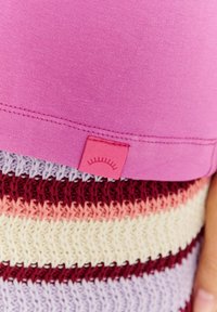 Růžový top s růžovou visačkou s jednoduchým designem, položený na vícero barevné pletenině se pruhy levandulové, růžové a burgundské barvy.