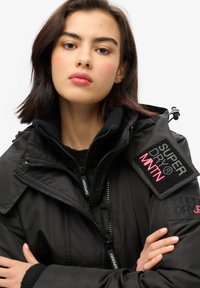 Czarna kurtka zewnętrzna z zamkiem błyskawicznym, wysokim kołnierzem i wieloma kieszeniami na zamek. Posiada naszywki z logo w różowym i czarnym kolorze na ramieniu.